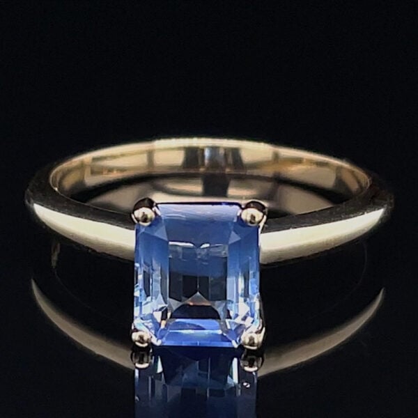 Emerald-cut blue gemstone ring on a silver band against a black background, shown in a close-up with reflection beneath the stone.