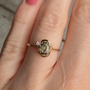 Gold ring with an oval pale-green gemstone and a small diamond, worn on a finger; delicate intertwined setting.