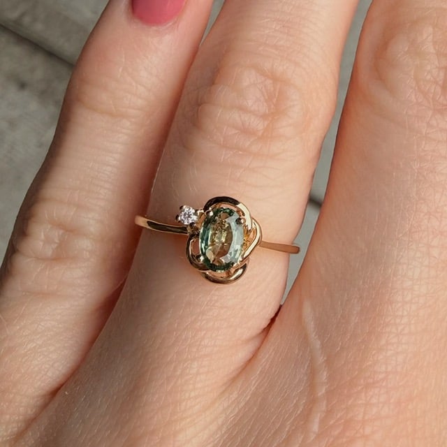 Gold ring with an oval pale-green gemstone and a small diamond, worn on a finger; delicate intertwined setting.