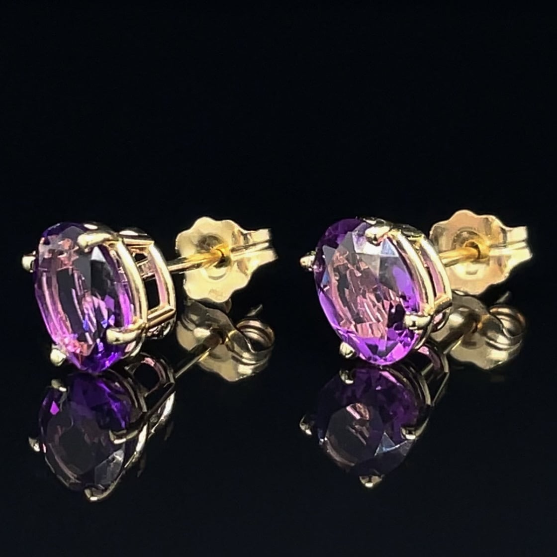 Pair of purple gemstone stud earrings in gold settings with butterfly backs, resting on a glossy black surface with reflections.