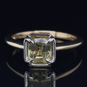 Ring with a pale yellow rectangular cushion-cut diamond in a four-prong setting on a dark background. Mention of a slim metal band (likely white gold).