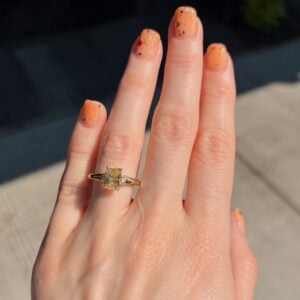 Hand with orange speckled nails wearing a gold ring featuring a yellow rectangular-cut gemstone on the ring finger, outdoors in sunlight.
