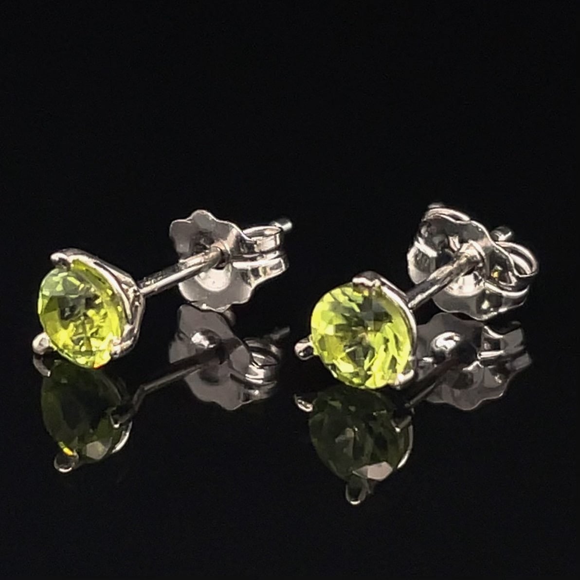 Pair of silver stud earrings, each with a bright lime-yellow peridot gemstone in a prong setting, against a black reflective background.