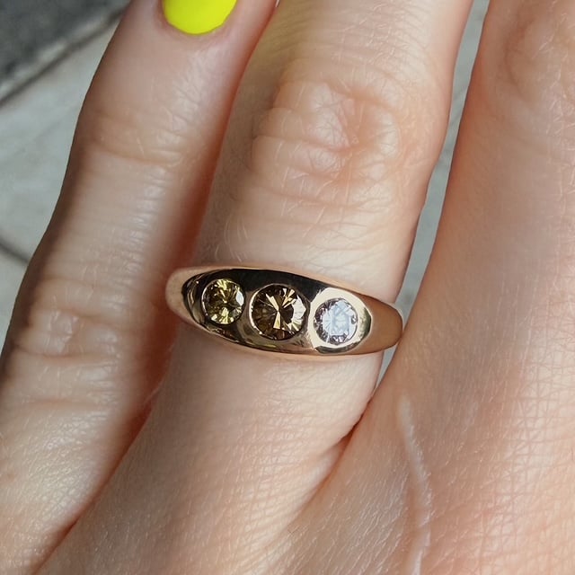 Gold ring with three round-cut stones (yellow, light brown, and clear) on a finger with neon-yellow nail polish nearby.