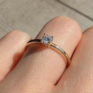 Close-up of a gold engagement ring with a round solitaire diamond on a finger outdoors.