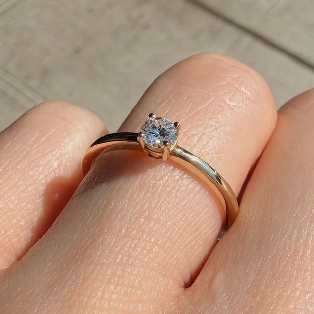 Close-up of a gold engagement ring with a round solitaire diamond on a finger outdoors.