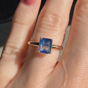 Ring on finger with a blue square-cut gemstone, gold band, close-up shot in bright light.