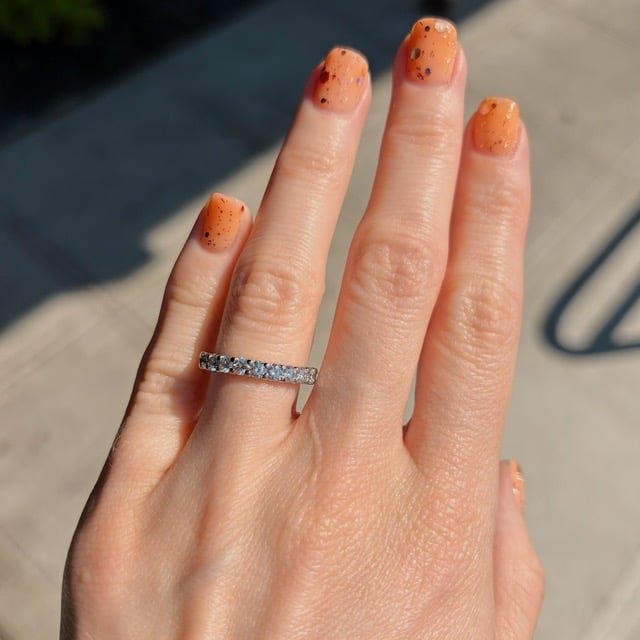 Hand with orange speckled nails wearing a diamond eternity ring on the ring finger.