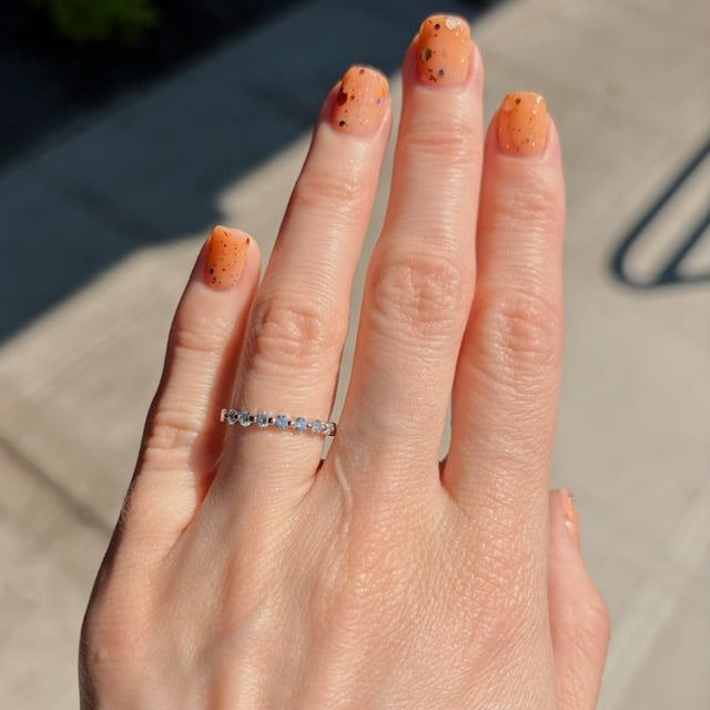 Hand with orange speckled manicure and a silver diamond-stone ring on the ring finger, outdoors.