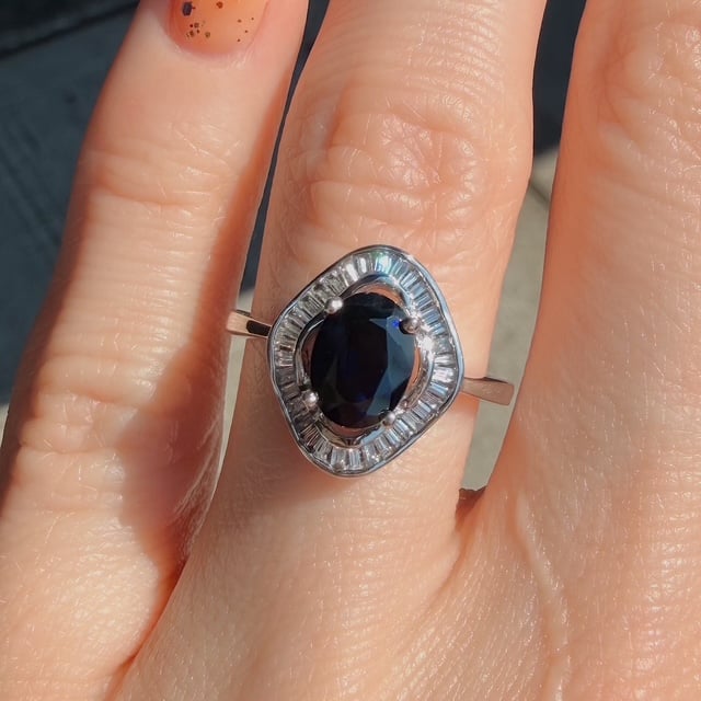 Ring with an oval dark blue gemstone surrounded by a halo of baguette-cut diamonds on a silver band, worn on a finger.