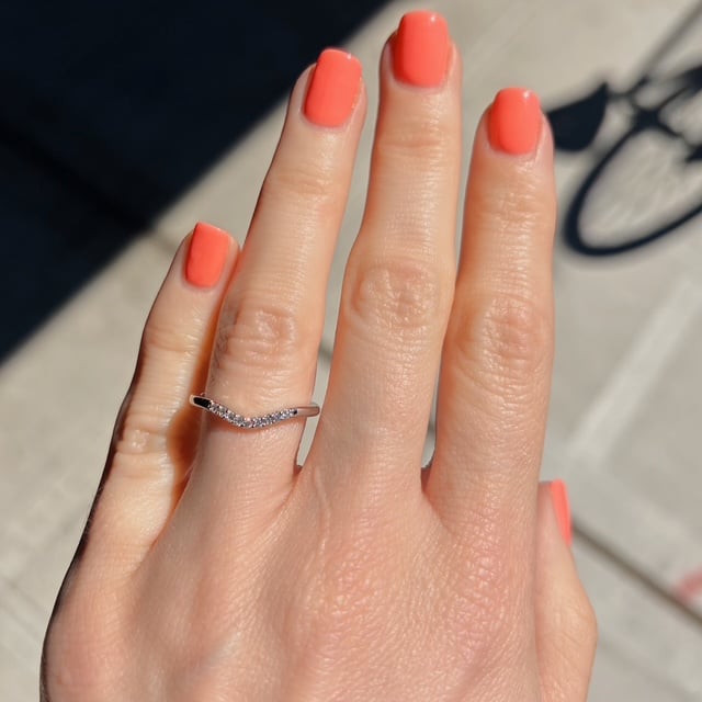 Hand with bright coral nail polish, wearing a slim wavy silver ring with small stones on the ring finger, outdoors.