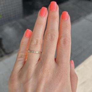Close-up of a hand with coral nail polish, wearing a thin gold ring with small square and round stones on the ring finger.
