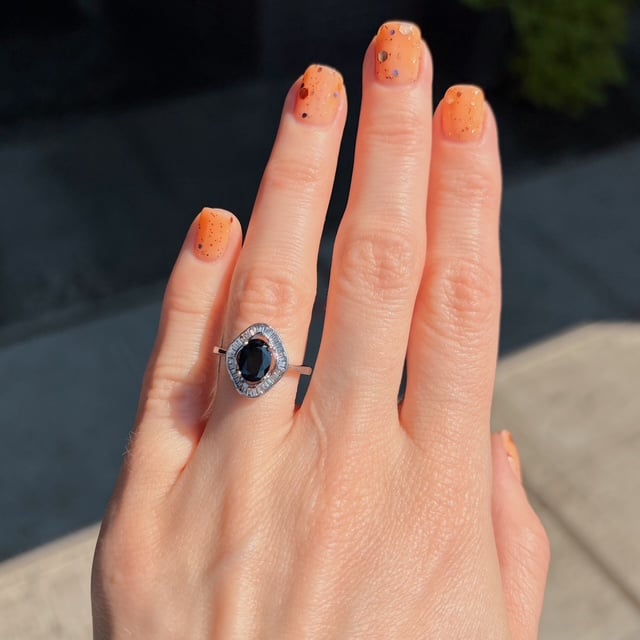 Hand with orange speckled nails wearing a ring with an oval black gemstone surrounded by a halo of small diamonds on the ring finger