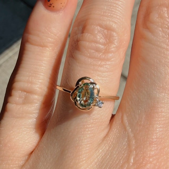 Close-up of a gold ring with an oval blue‑green gemstone and a small diamond, worn on a finger in sunlight.