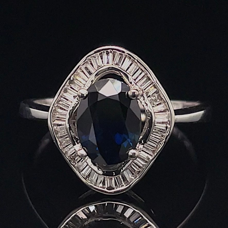 Silver ring with a dark blue oval cushion-cut center stone, surrounded by a halo of baguette diamonds on a black background.