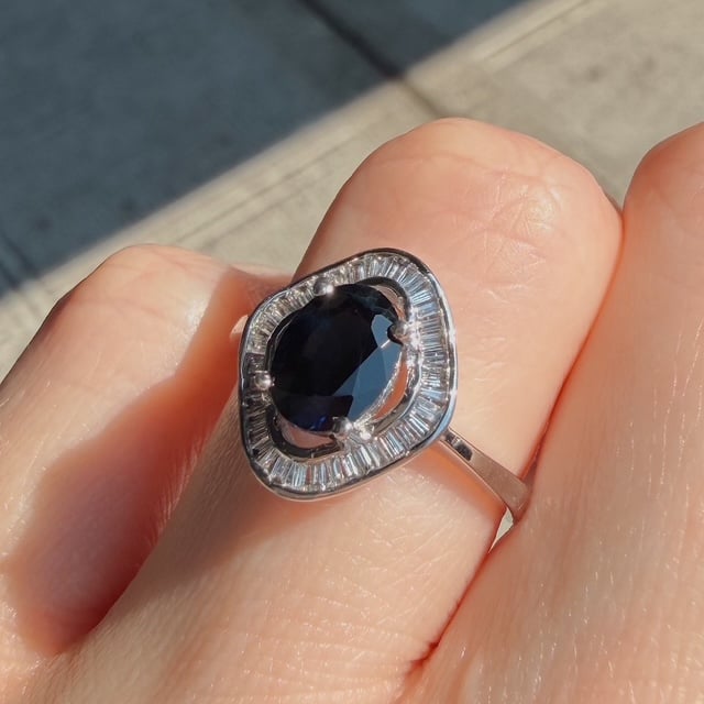 Close-up of a silver ring on a finger featuring a heart-shaped halo with a dark blue sapphire and surrounding baguette diamonds.