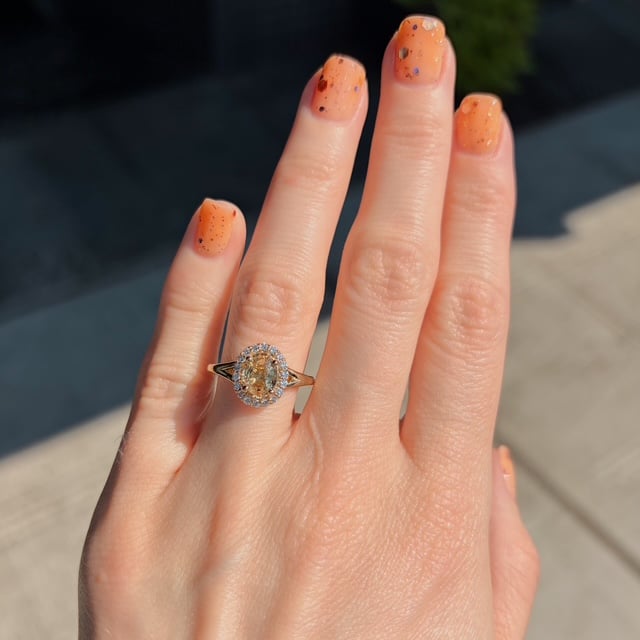 Close-up of a hand with orange nail polish wearing an oval halo engagement ring on the ring finger.