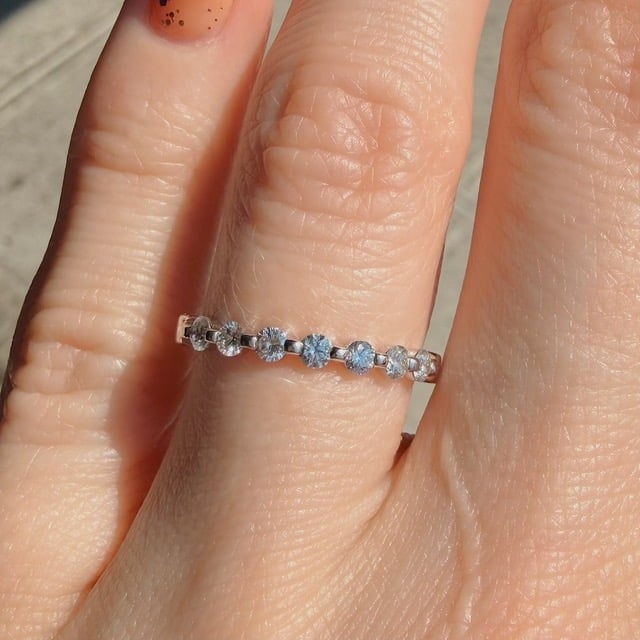 Close-up of a slim silver ring with a row of blue round-cut stones on a finger, sparkling in sunlight.