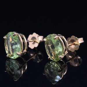 Oval pale-green gemstone stud earrings set in silver with butterfly backs, on a reflective black surface.