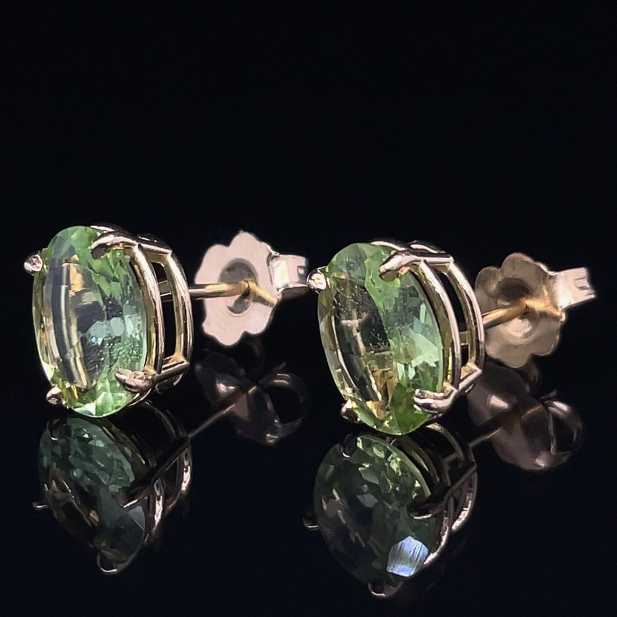Oval pale-green gemstone stud earrings set in silver with butterfly backs, on a reflective black surface.