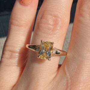 Gold ring with a rectangular pale yellow gemstone set in a prong setting, flanked by small diamonds on a finger.