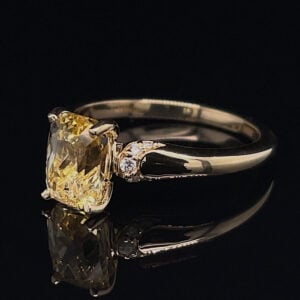 Engagement ring with a large cushion-cut yellow diamond and two small side diamonds on a polished white metal band, reflected on a black surface.