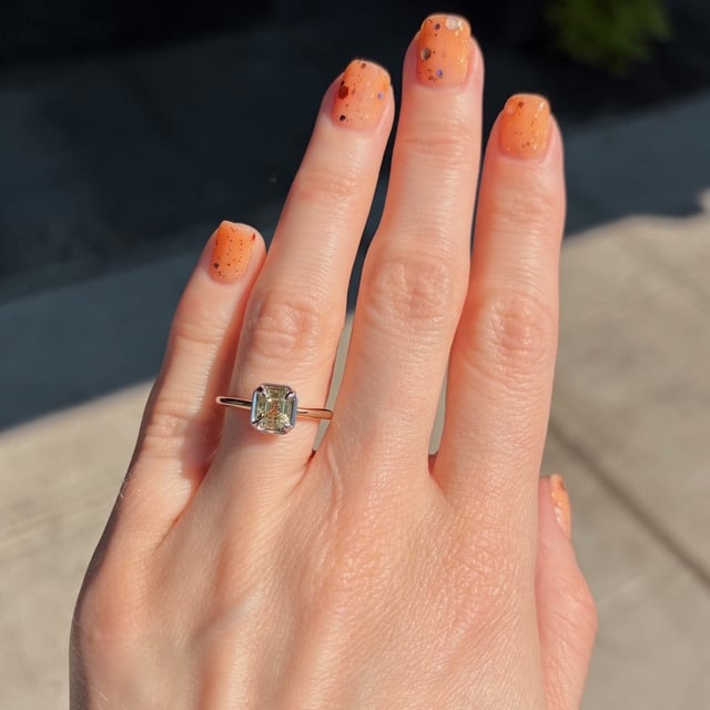 Hand with orange speckled nails wearing a gold ring with a square-cut diamond on the ring finger, outdoors