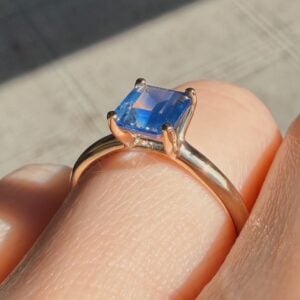 Close-up of a gold ring with a square-cut blue gemstone resting on a finger in sunlight.