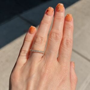 Close-up of a hand with orange speckled nail polish and a diamond-studded band on the ring finger.