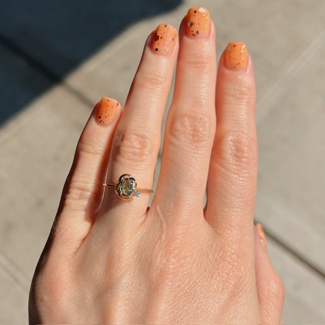 Left hand with orange speckled nails wearing a gold ring with an oval blue-green gemstone on the ring finger outdoors.