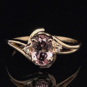 Gold ring with an oval pink center stone and small side diamonds on a curved band against a black background, close-up.
