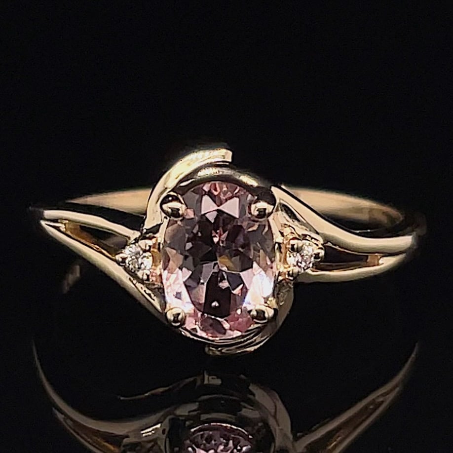 Gold ring with an oval pink center stone and small side diamonds on a curved band against a black background, close-up.
