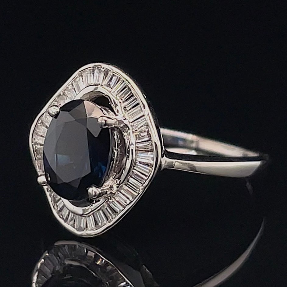 Oval black/dark blue gemstone ring with a halo of rectangular clear stones set in white metal.