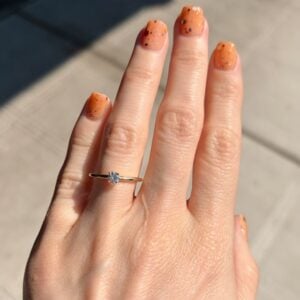 Close-up of a left hand showing orange speckled nails and a gold ring with a single diamond on the ring finger, outdoors. (sunny day)