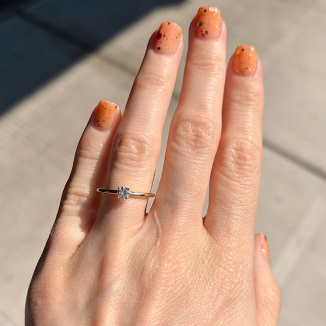 Close-up of a left hand showing orange speckled nails and a gold ring with a single diamond on the ring finger, outdoors. (sunny day)