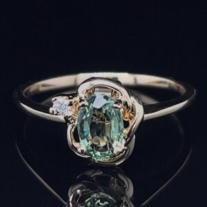 Oval green gemstone ring with a small diamond accent on a two-tone band, shown on a dark reflective surface.