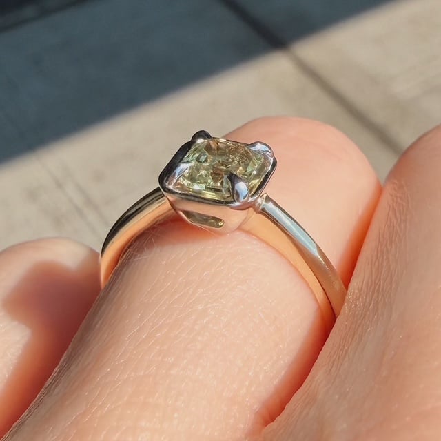 Engagement ring with a pale yellow rectangular diamond on a silver band worn on a finger in sunlight, close-up view.