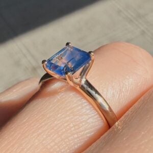 Rose gold ring featuring a square-cut blue gemstone held by four prongs on a finger, with sunlight highlighting the facets.