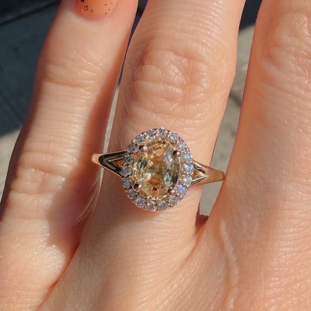 Gold ring featuring an oval yellow gemstone surrounded by a halo of small diamonds on a finger.