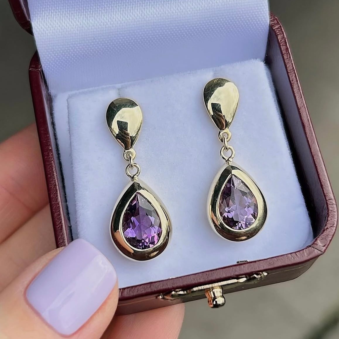 Gold teardrop earrings with lavender-purple stones in a blue-lined jewelry box.