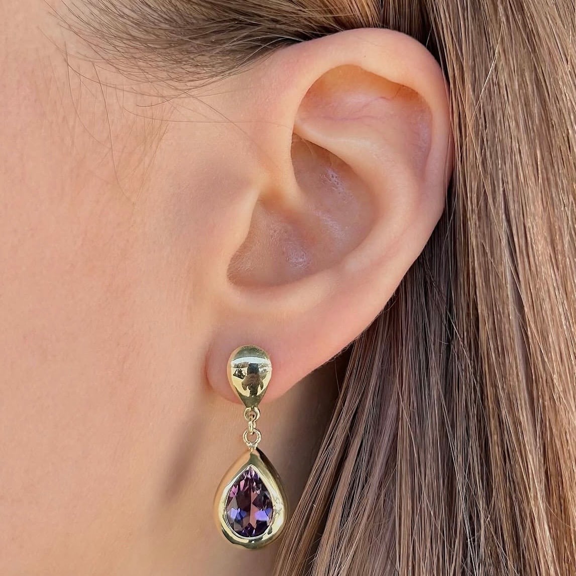 Gold teardrop earring with purple gem hanging from a stud on an ear lobe.