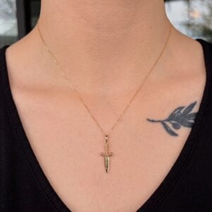 Gold necklace with a dagger pendant resting on the chest, with a leaf tattoo visible on the collarbone area.