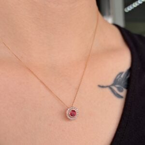 Rose gold necklace with a red central gemstone surrounded by a halo of small diamonds resting on the collarbone; tattoo visible on the shoulder.
