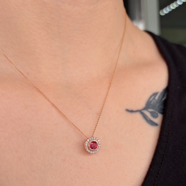 Rose gold necklace with a red central gemstone surrounded by a halo of small diamonds resting on the collarbone; tattoo visible on the shoulder.