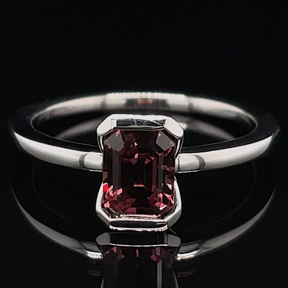 Close-up of a silver ring with a rectangular pink gemstone centered on a black reflective background.