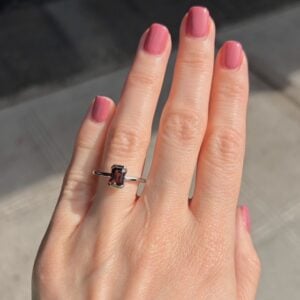 Close-up of a hand with pink nails wearing a silver ring with a rectangular pink gemstone on the ring finger.
