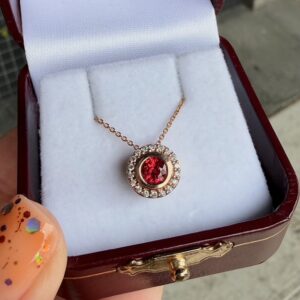 Gold pendant with a red center gemstone surrounded by a halo of small clear stones in a white padded jewelry box.