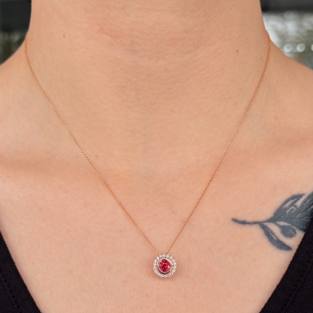 Gold chain necklace with a circular pendant featuring a red central gem surrounded by clear stones on a bare neck with a partial tattoo nearby.