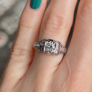 Close-up of a silver vintage ring on a finger with teal nail polish, featuring a square diamond cluster centerpiece and decorative engravings on the band
